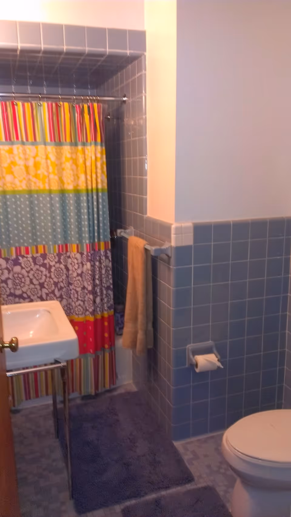 Small tiled bathroom with a colorful striped and floral shower curtain, pedestal sink, toilet, towel on a rack, and blue bath mats.