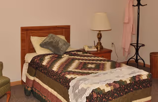 A single bed with a multicolored patchwork quilt and pillows in a room. Next to the bed is a wooden nightstand with a brass lamp and a telephone. A coat rack with a pink robe hanging on it and a green upholstered chair are also visible.