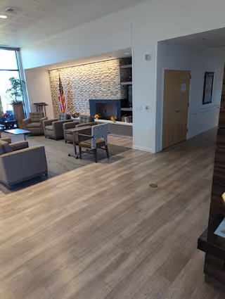 A spacious interior common area with light wood flooring, several gray armchairs arranged around a modern stone fireplace, an American flag in the corner, and large windows letting in natural light.