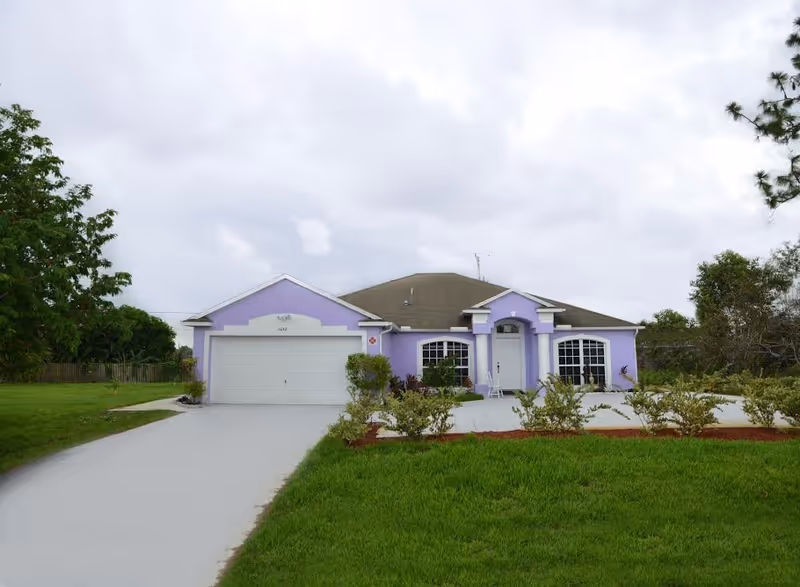 Single-story lavender house with an attached garage, front lawn, and driveway under a cloudy sky.