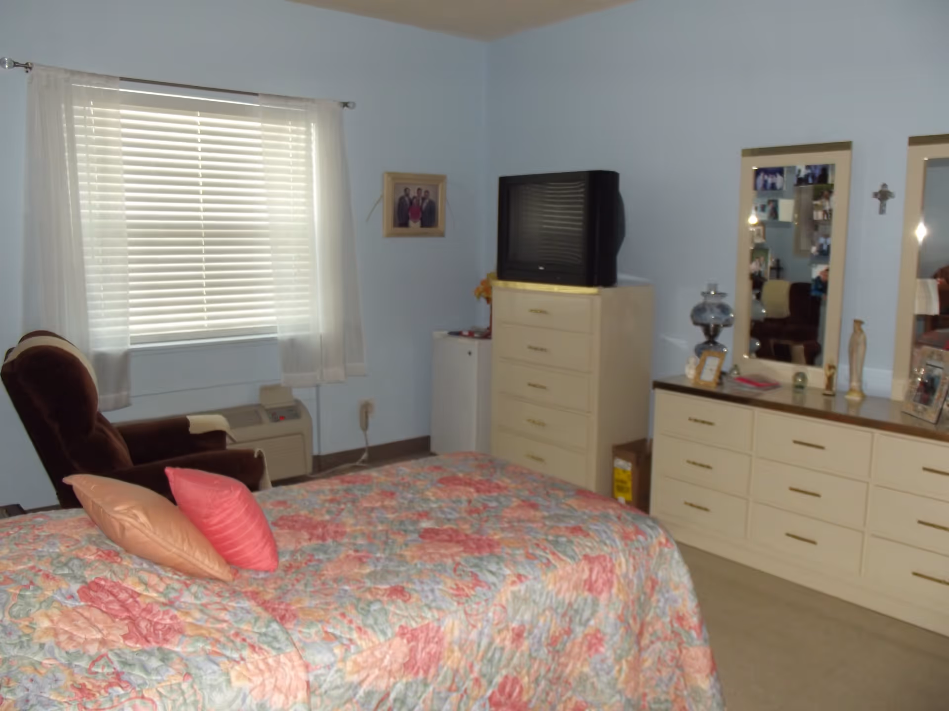 A bedroom with a bed covered in a floral quilt and two pillows, a brown armchair near a window with white blinds and sheer curtains, a dresser with a TV on top, and a long dresser with two mirrors and various decorative items on it.