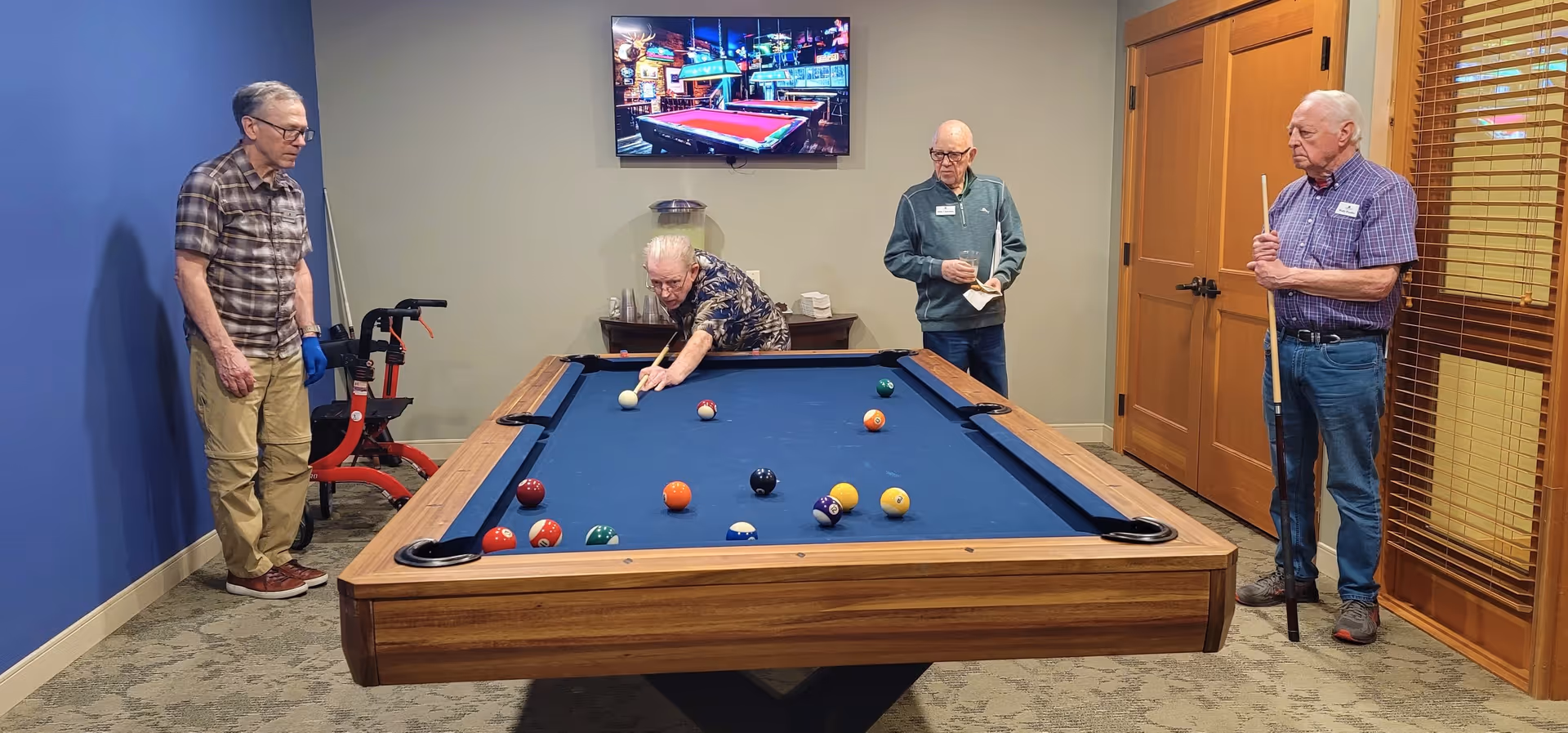 Four elderly men playing pool in a game room with a blue pool table. One man is taking a shot while the others watch. The room has a blue wall on the left, beige walls, a carpeted floor, wooden doors, and a TV mounted on the wall showing a pool hall scene.