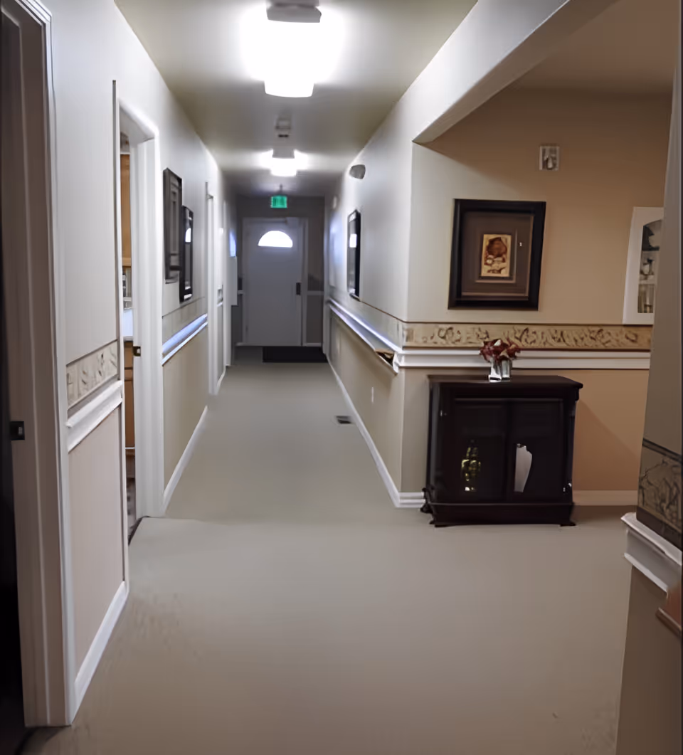 A well-lit hallway in an assisted living facility with beige walls and carpeted floor. The hallway has handrails on both sides, framed artwork on the walls, and a small dark wooden cabinet with a vase of flowers on top. At the end of the hallway is a door with a window and an illuminated exit sign above it.