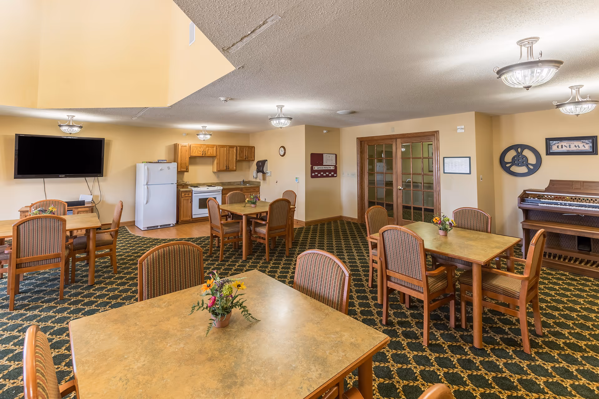 A communal dining and activity room with several tables and chairs arranged on a patterned carpet. There is a small kitchen area with a refrigerator, stove, and cabinets in the background. A large flat-screen TV is mounted on the wall to the left. On the right side, there is a wooden organ and wall decorations including a film reel and a sign that says 'Home Cinema'. The room is well-lit with ceiling lights and has a warm, inviting atmosphere.