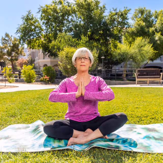An elderly woman with short gray hair and glasses is sitting cross-legged on a yoga mat on a grassy lawn outdoors. She is wearing a long-sleeve purple top and black pants, with her hands pressed together in a prayer position at chest level. Behind her are trees, shrubs, benches, and a building under a clear blue sky.