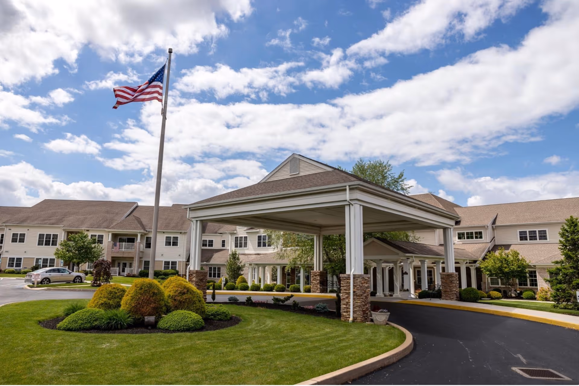Front exterior of Homewood Living Plum Creek with a covered porte-cochère, landscaped grounds, and an American flag under a partly cloudy sky.