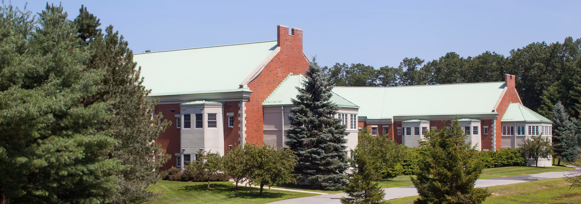 Exterior view of a large brick building with a light green roof surrounded by trees and greenery under a clear blue sky.