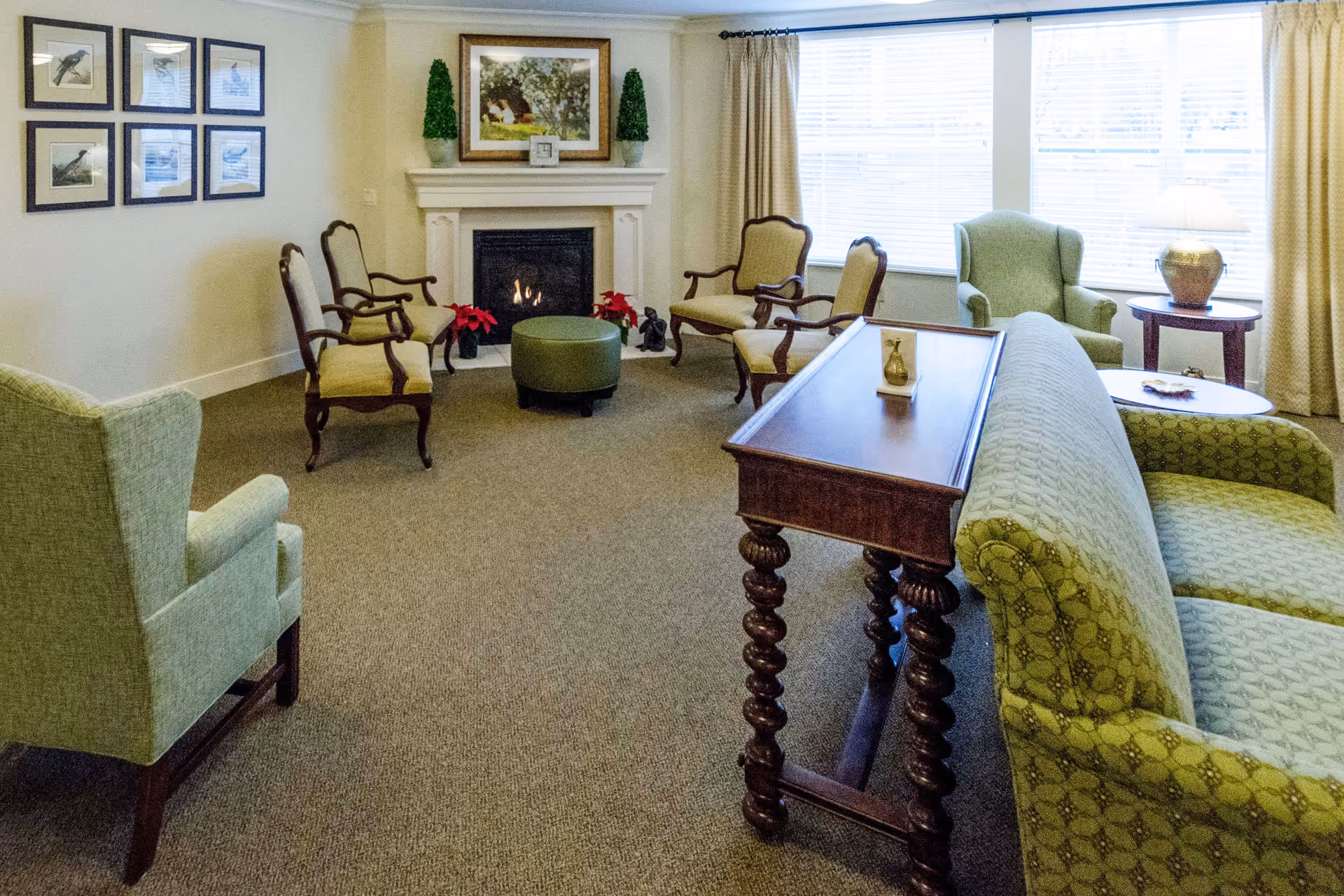 A cozy senior living room with a green patterned sofa, two green armchairs, and four wooden chairs with beige cushions arranged around a lit fireplace. A wooden table with ornate legs is placed behind the sofa. The room has a large window with beige curtains, a side table with a lamp, framed bird pictures on one wall, and decorative plants on the fireplace mantel.