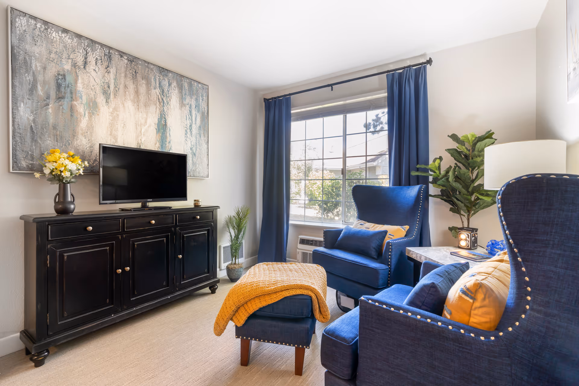 A cozy living room with two blue armchairs and a matching ottoman covered with a yellow throw blanket. A black cabinet holds a flat-screen TV and a vase with yellow and white flowers. A large abstract painting hangs above the cabinet. The room has a large window with blue curtains, a side table with a lamp, and a potted plant in the corner.
