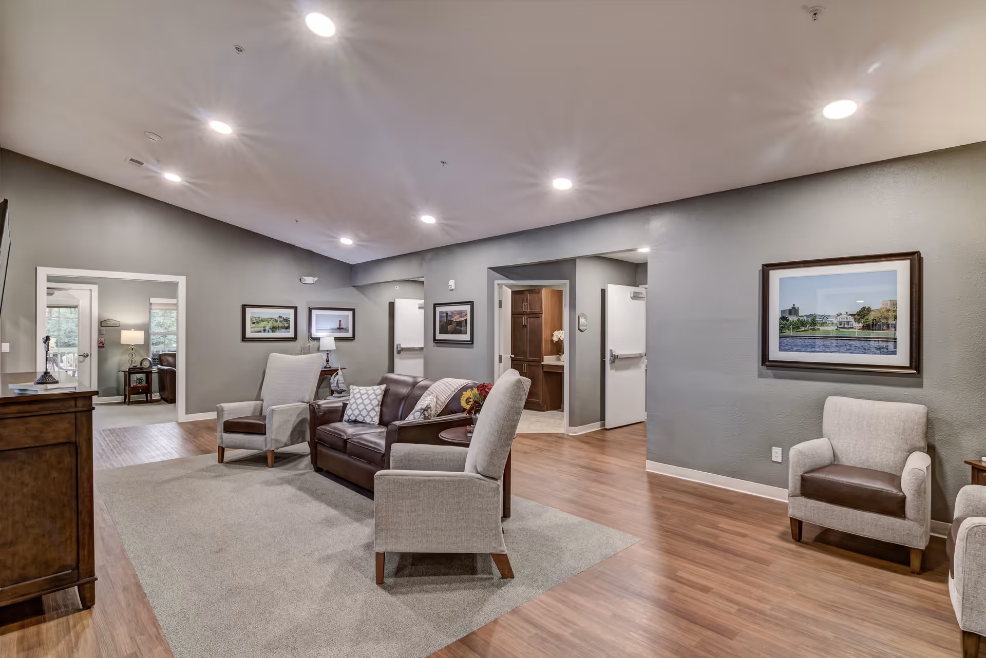 Bright common lounge with sofas and armchairs arranged on a rug, framed artwork on gray walls, and wood floors.