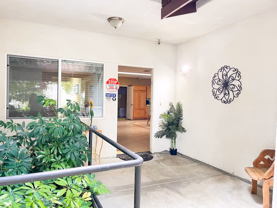Entrance area of a facility with a doorway leading inside. There are green plants on the left side and a potted plant on the right. A decorative metal wall art is mounted on the white wall to the right. A wooden chair is partially visible on the right side. The floor is concrete and the interior beyond the doorway has tiled flooring and wooden cabinets.