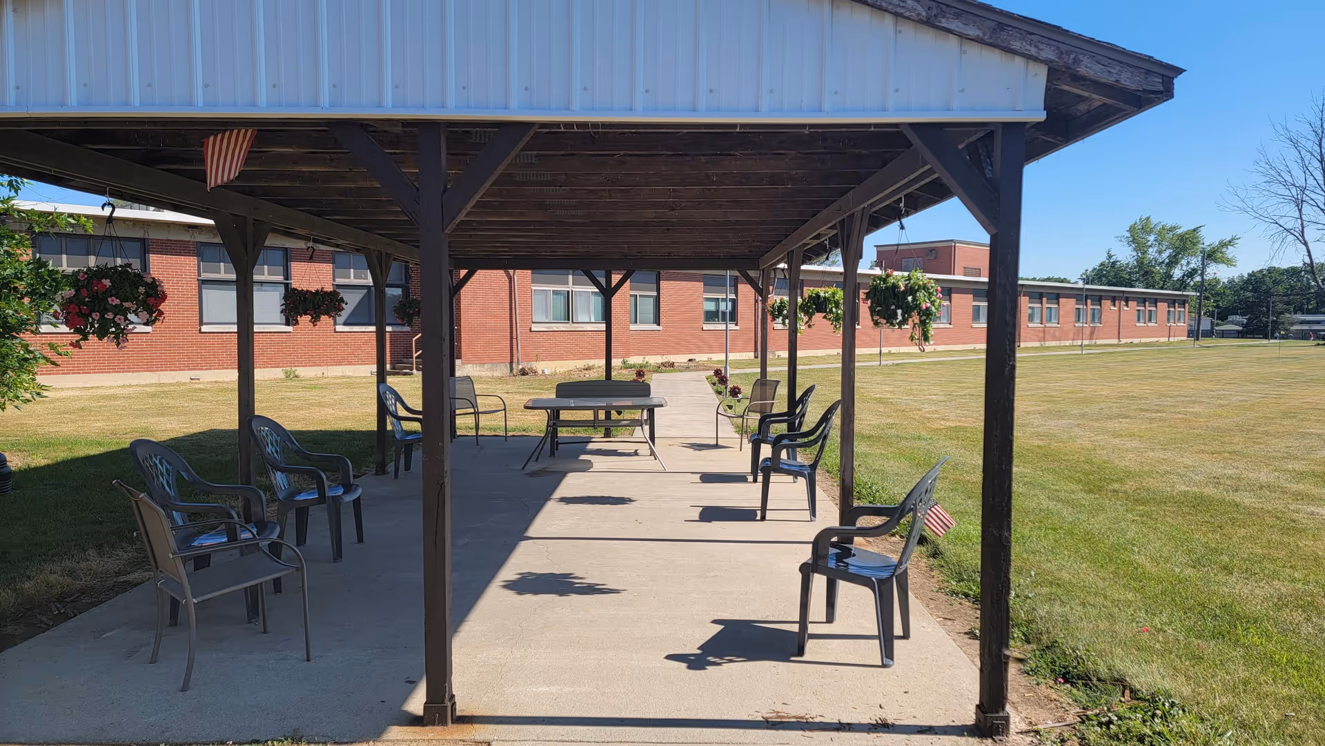 Outdoor covered seating area with several plastic chairs and a table under a wooden pavilion. Hanging flower baskets are attached to the pavilion beams. In the background, there is a large brick building and a well-maintained grassy lawn under a clear blue sky.