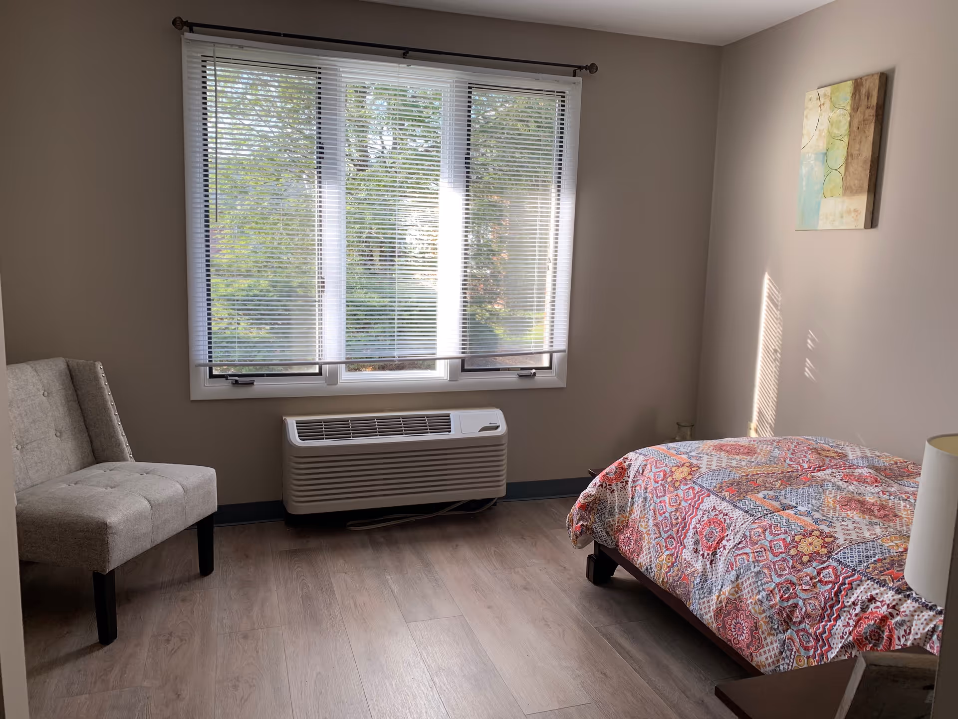 A simple bedroom with a large window covered by white blinds, a beige upholstered chair, a bed with a colorful patterned quilt, a small nightstand with a lamp, and a piece of abstract artwork on the wall.