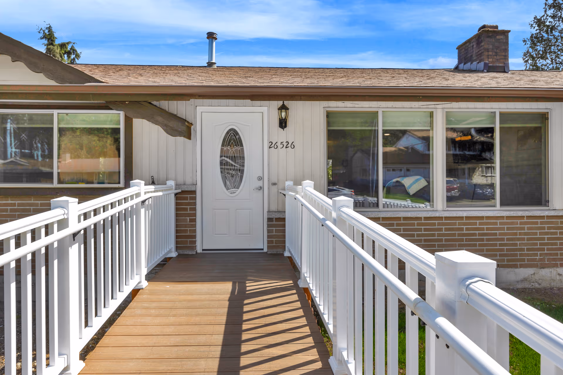 Front entrance of a single-story building with a white door featuring an oval glass design, flanked by large windows. A wooden ramp with white railings leads up to the door. The building has a brick lower exterior and light-colored siding above. The address number 26526 is displayed next to the door under a wall-mounted light fixture.
