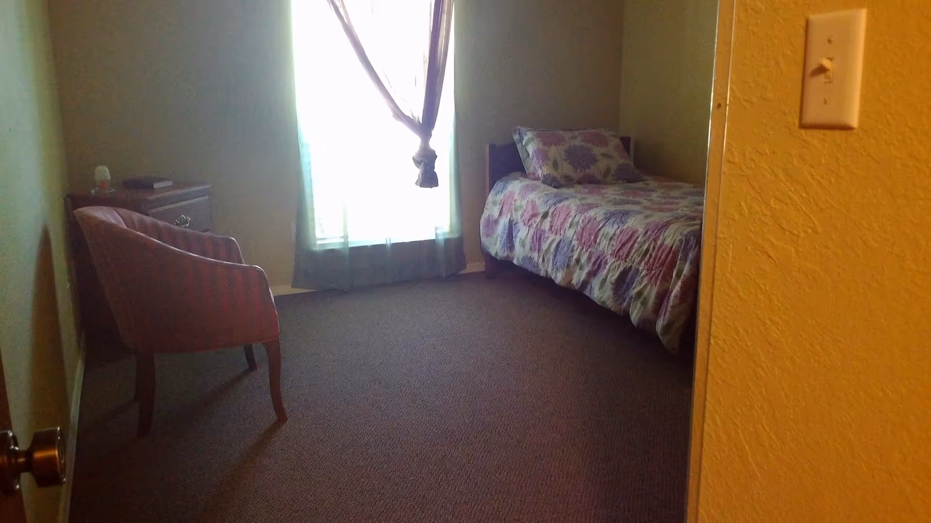 A small bedroom with a single bed covered in a floral quilt, a wooden nightstand with items on it, a striped upholstered chair, and a window with sheer curtains tied in the middle letting in light.
