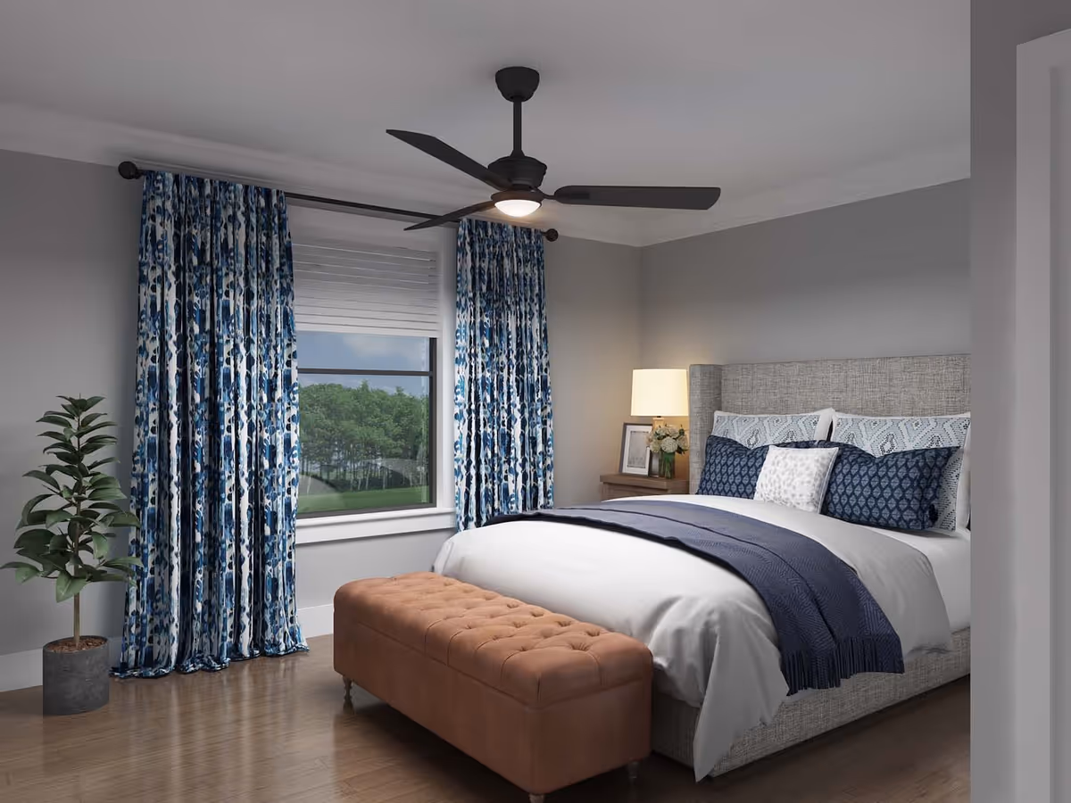 A modern bedroom with a large bed featuring a gray upholstered headboard, white and blue bedding, and multiple pillows. At the foot of the bed is a tufted brown bench. To the left, there is a window with blue patterned curtains and a white blind, showing a view of green trees outside. A potted plant is placed near the window. On the right side of the bed, there is a wooden nightstand with a lamp, a framed picture, and a small flower arrangement. The room has gray walls, wooden flooring, and a ceiling fan with a light.