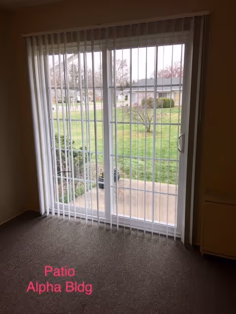 View through vertical blinds covering a sliding glass door that opens to a small concrete patio and a grassy outdoor area with trees and buildings in the background.