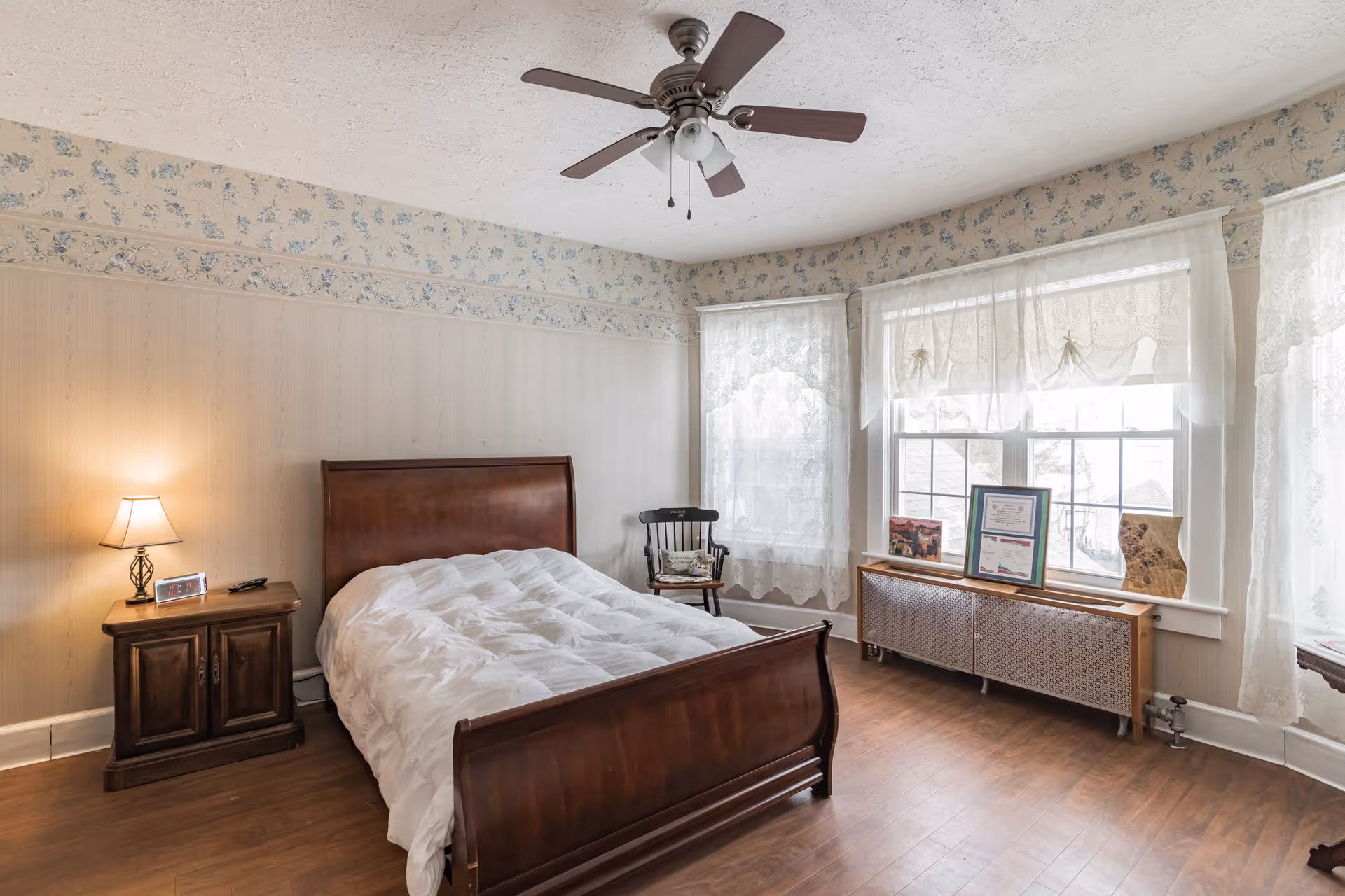 A cozy bedroom with a wooden bed frame and white comforter, a wooden nightstand with a lamp and digital clock, a wooden chair near two windows with lace curtains, and a wooden radiator cover with framed pictures and certificates on top. The room has floral wallpaper and a ceiling fan with lights.