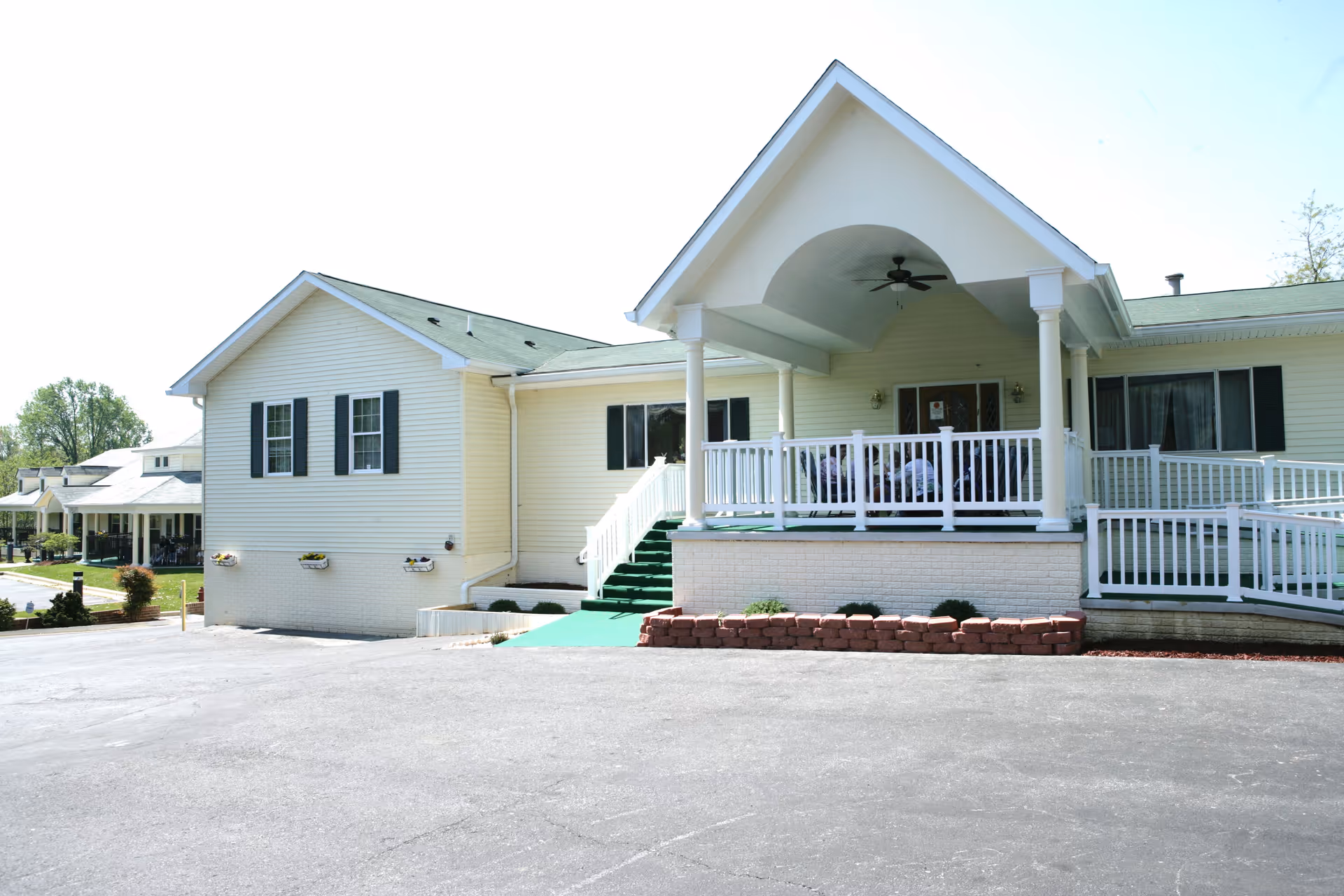 Front view of a single-story light-yellow senior living building with a covered porch, white railings, ramp, and steps.