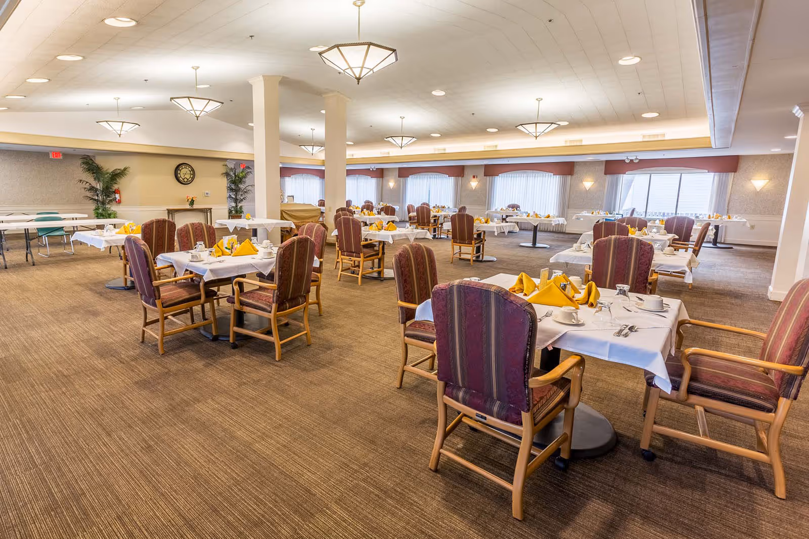 A spacious dining room with multiple tables covered in white tablecloths and set with yellow folded napkins, cups, and utensils. The room has carpeted floors, large windows with sheer curtains, and several ceiling lights. There are upholstered wooden chairs around each table and some potted plants along the walls.