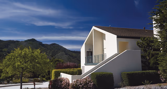 White two-story building with an exterior staircase and balcony set among landscaped shrubs and trees with green hills and a blue sky in the background.