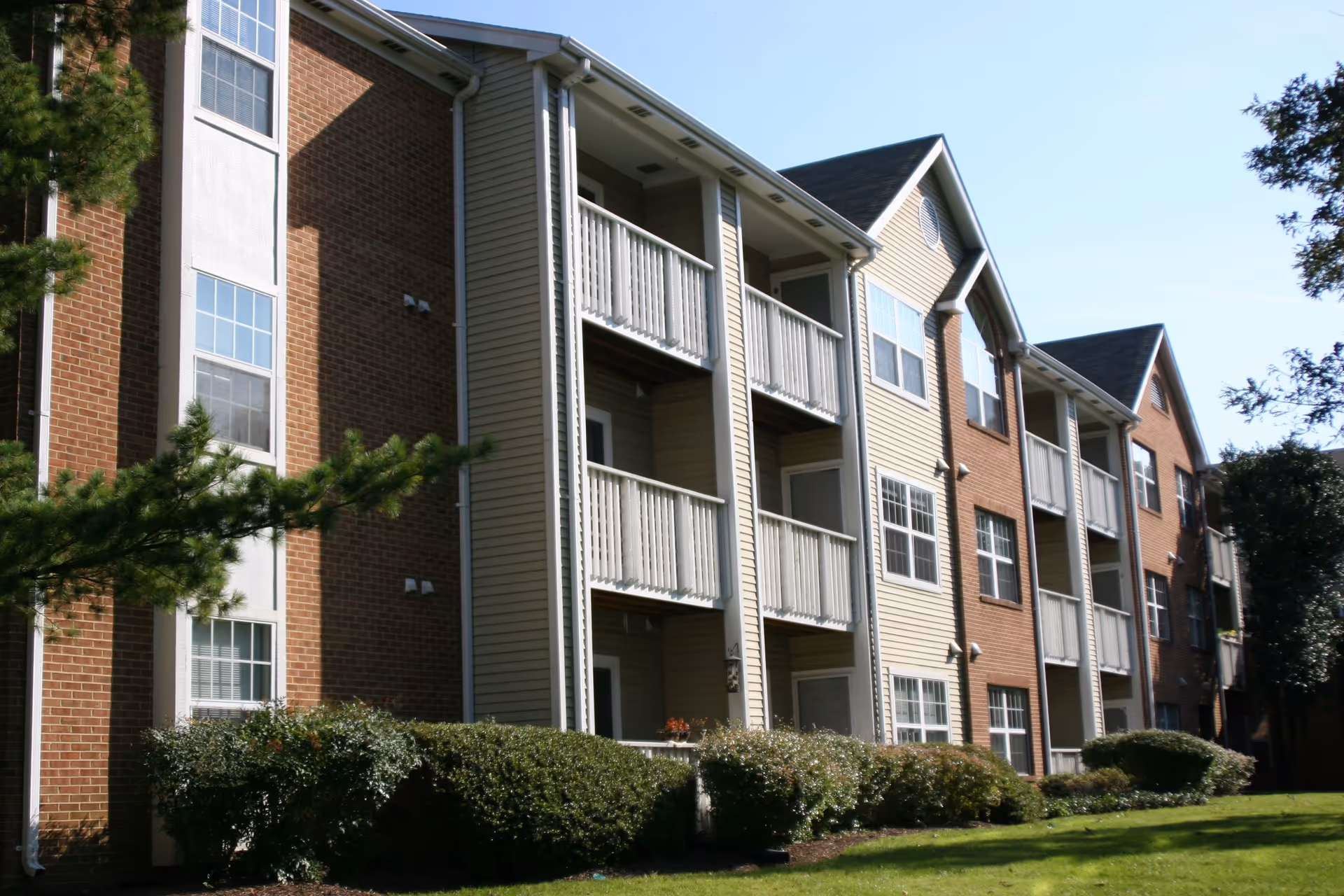 Exterior view of a multi-story residential building with brick and siding walls, multiple balconies, and several windows. The building is surrounded by green bushes and a well-maintained lawn under a clear blue sky.