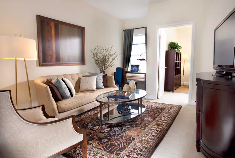 A cozy living room with a beige sofa adorned with multiple pillows, a glass coffee table with decorative items and books, a patterned area rug, a beige armchair, a floor lamp, and a large framed artwork on the wall. In the background, there is a small desk with a laptop and a chair near a window, and an open doorway leading to another room with a dark wooden cabinet and a floor lamp.