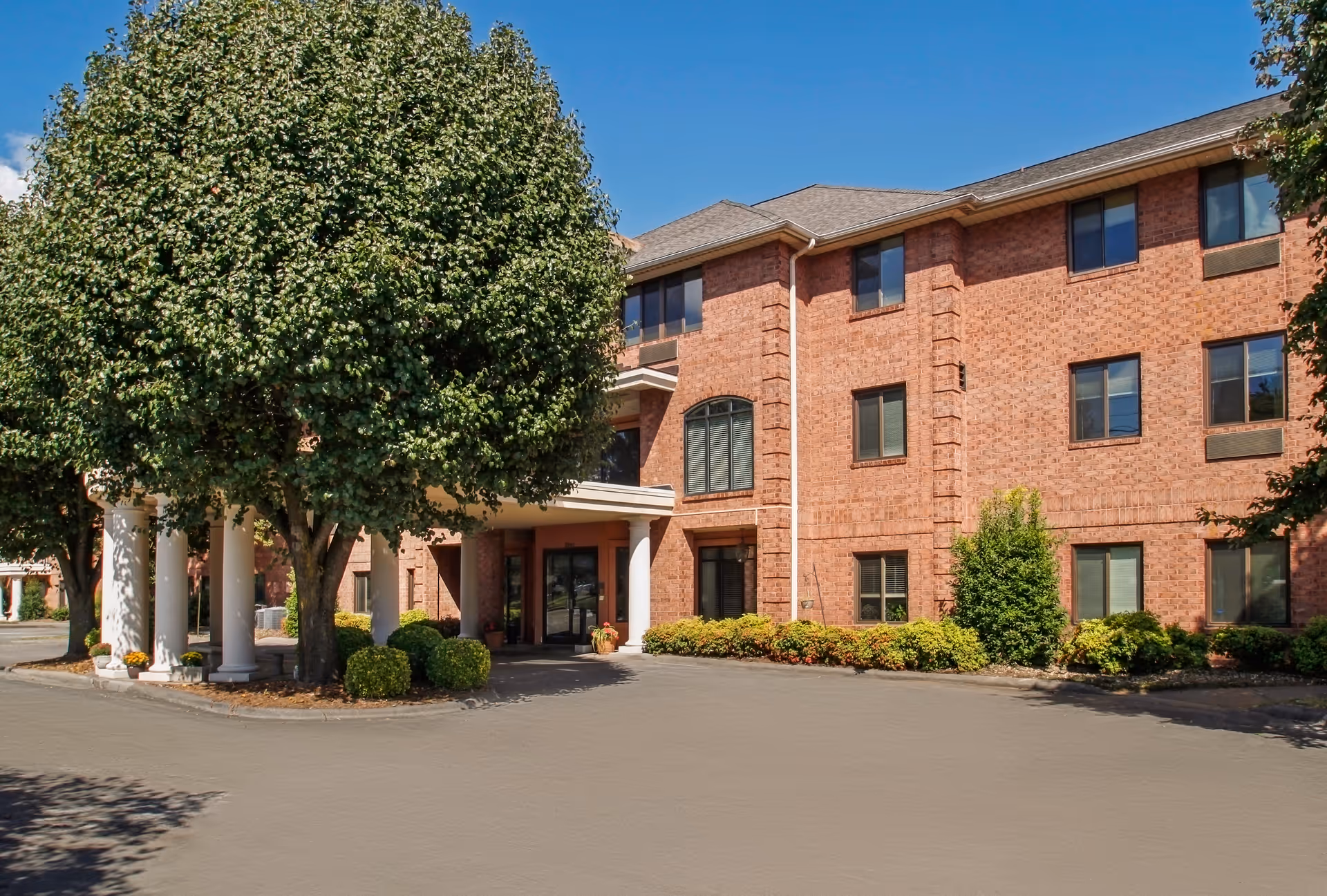 Brick three-story senior living facility entrance with columns, trees, and a driveway.