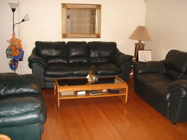 A living room with three dark green leather sofas arranged around a wooden coffee table. The coffee table has a glass top and some decorative items underneath. There is a floor lamp with two lights on the left side and a table lamp with a beige shade on the right side. A framed mirror hangs on the wall above the central sofa. The floor is wooden.