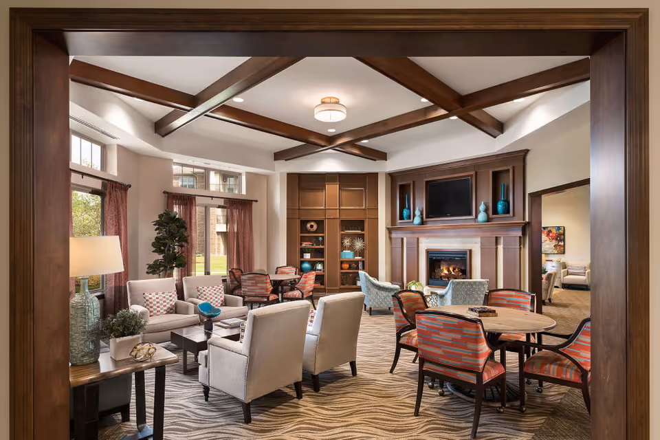 A cozy living room area in Cedarbrook of Northville featuring a fireplace with a mounted TV above it, wooden ceiling beams, multiple seating arrangements including armchairs and sofas with patterned cushions, a round table with chairs, large windows with curtains allowing natural light, and decorative vases on shelves and mantel.