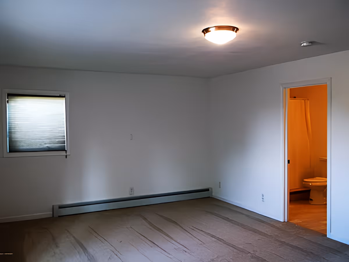 Empty room with beige carpet, white walls, a small window with a closed blind, and a ceiling light. There is an open doorway leading to a bathroom with a visible toilet and shower curtain.