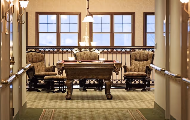 A hallway leading to a recreational area with a pool table and three cushioned armchairs. The area is carpeted and well-lit with hanging pendant lights and wall sconces. Large windows in the background let in natural light.