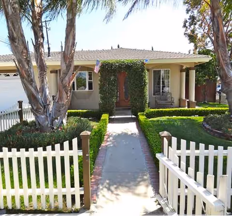 Front view of a single-story senior living home with a walkway, ivy arch, and white fence.