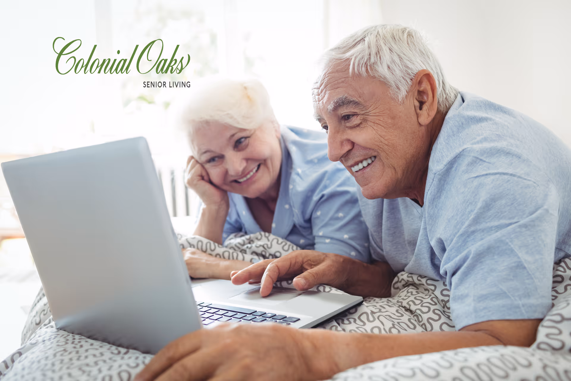 An elderly couple lying in bed together, smiling and looking at a laptop screen. The woman has white hair and is resting her head on her hand, while the man is using the laptop. They are covered with a patterned blanket in a bright, softly lit room.