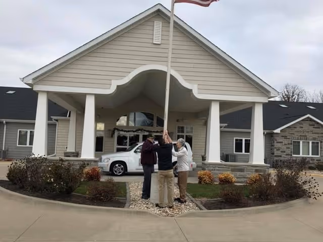 Three people standing together holding a flagpole in front of the entrance to a single-story building with a covered porch and a white vehicle parked nearby. The building has beige siding and stone accents with landscaped bushes around the entrance.