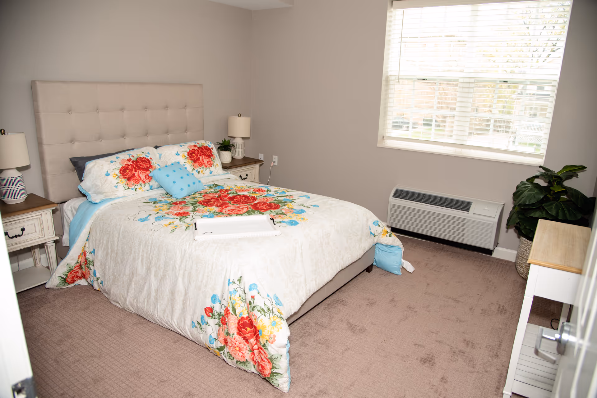A bedroom with a beige tufted headboard bed covered with a floral comforter featuring red, blue, and yellow flowers. There are two matching floral pillows and a small blue decorative pillow on the bed. On either side of the bed are white nightstands with table lamps. A window with white blinds is on the right wall, and below it is a wall-mounted air conditioning unit. A green potted plant is placed near the window, and a small white table is positioned near the door.