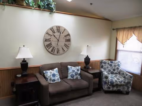 Cozy living room lounge with a brown sofa, patterned chair, lamps, and a large wall clock.