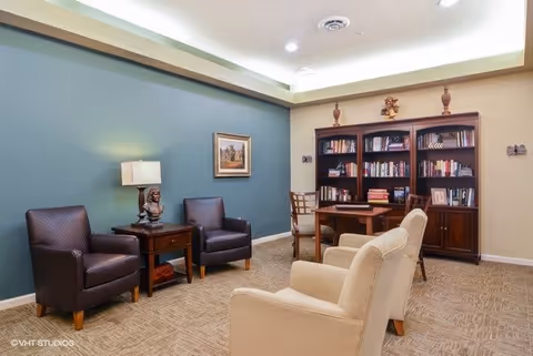 A cozy room with a blue accent wall featuring a framed painting, two dark leather armchairs with a wooden side table and lamp between them, two beige upholstered chairs facing a wooden desk with a chair, and a large wooden bookshelf filled with books and decorative items.