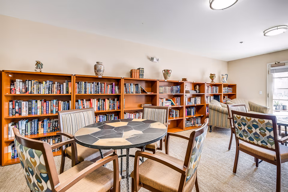 A bright and cozy reading room with wooden bookshelves filled with books along the wall. There are several chairs with patterned upholstery around a round table with a stone tile top. Two armchairs are positioned near a glass door that leads outside, allowing natural light to fill the room.