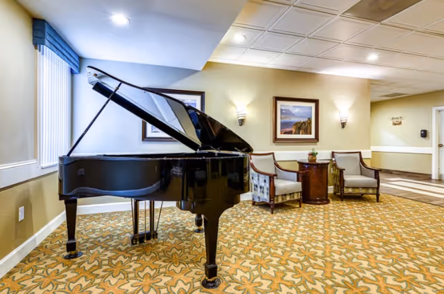 A black grand piano in a carpeted sitting area with upholstered chairs, a side table, and framed wall art.