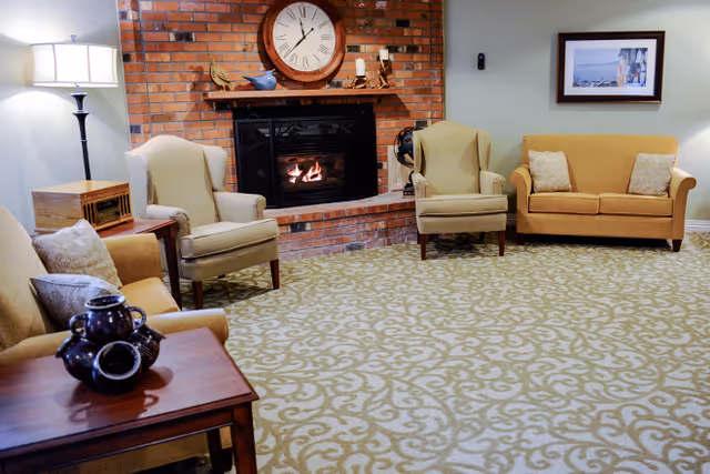 A cozy living room with a brick fireplace in the center, a large clock above the fireplace, two beige armchairs, a beige sofa with two pillows, a wooden side table with decorative items, a floor lamp, and a framed picture on the wall.