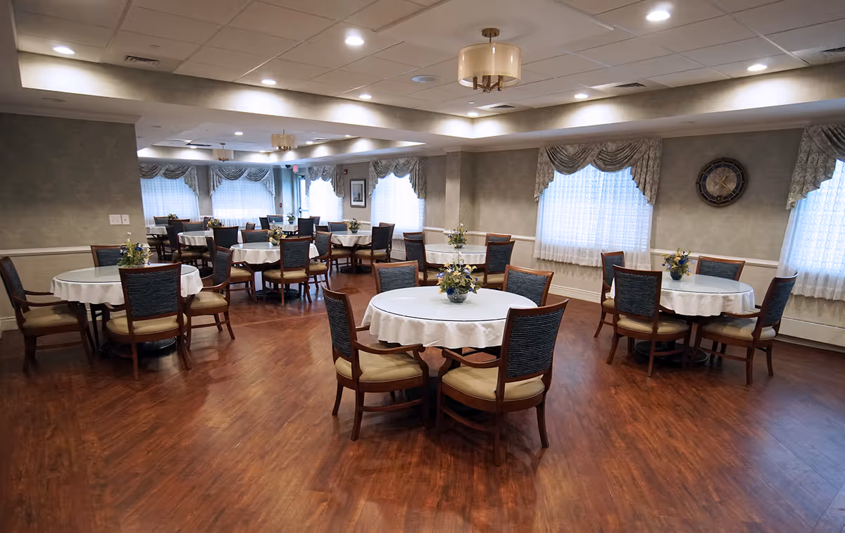 Spacious dining room with round tables covered in white tablecloths, wooden chairs, floral centerpieces, and large curtained windows.