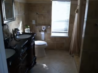 A bathroom with beige tiled walls and floor, featuring a dark wooden vanity with a sink and mirror, a toilet next to a window with blinds, and a shower area with a curtain.