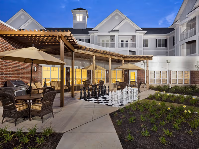 Outdoor patio area at Irene Woods Assisted Living featuring wicker chairs with cushions, tables with large umbrellas, a pergola, and a large outdoor chess set on a checkered board. The building exterior is visible in the background with lit windows and a clear evening sky.