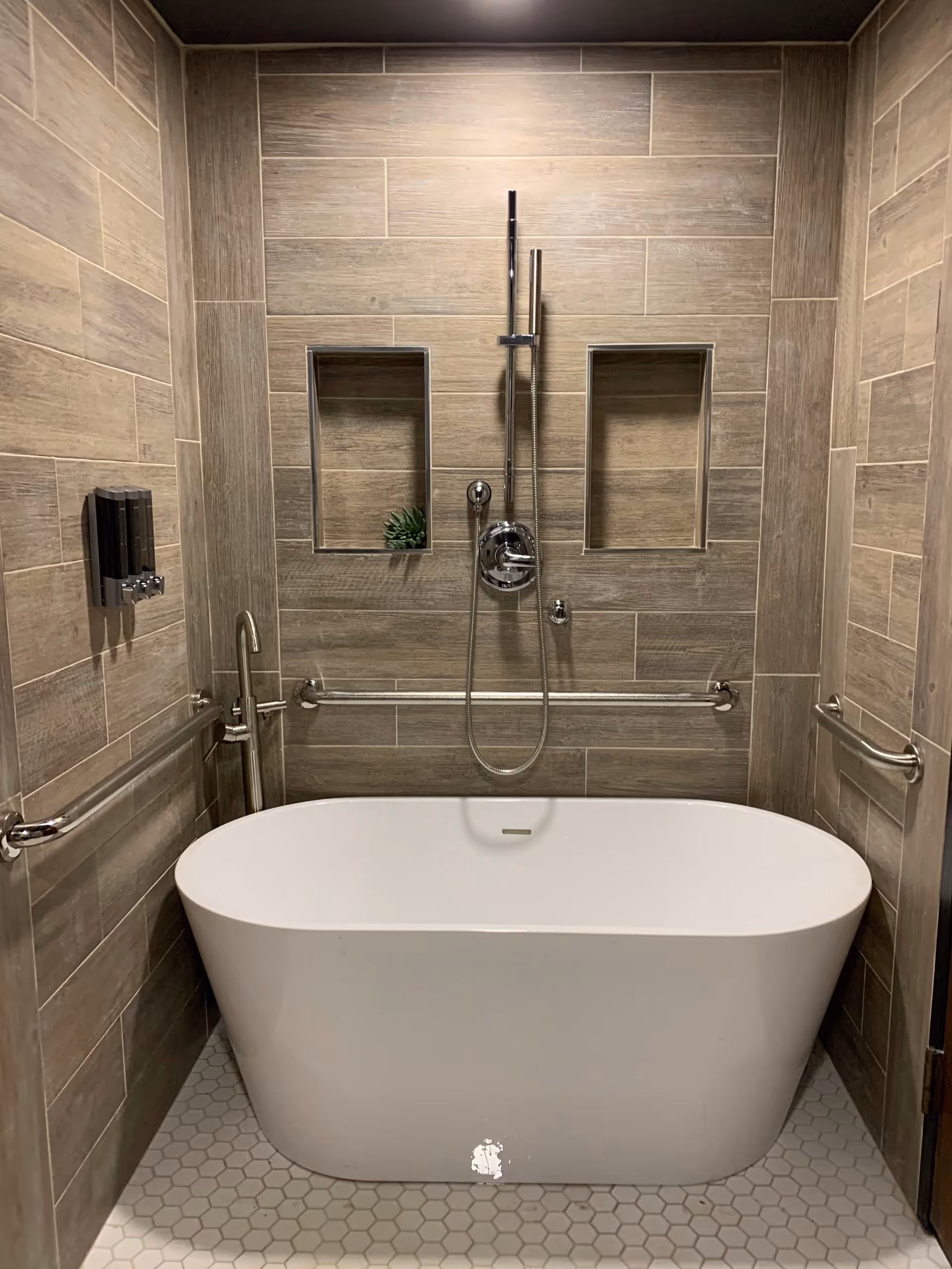 Modern tiled bathroom featuring a freestanding white bathtub, wall-mounted showerhead and handheld, grab bars, and two recessed shelves.