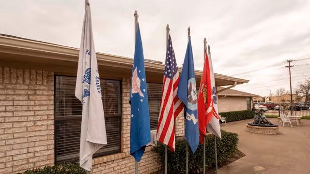 Five flags displayed on poles outside a brick building with windows and bushes. The flags include the United States flag and various military or organizational flags. There is a small circular brick structure with a statue or sculpture and some outdoor seating visible in the background under a cloudy sky.