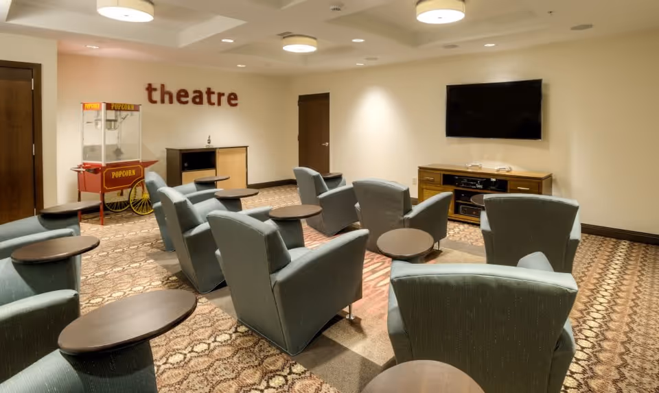 A small theater room with multiple green upholstered armchairs arranged in rows facing a wall-mounted flat screen TV. Each chair has a small attached round table. There is a popcorn machine on the left side of the room and the word 'theatre' displayed on the wall above a cabinet. The room has patterned carpet and soft ceiling lights.