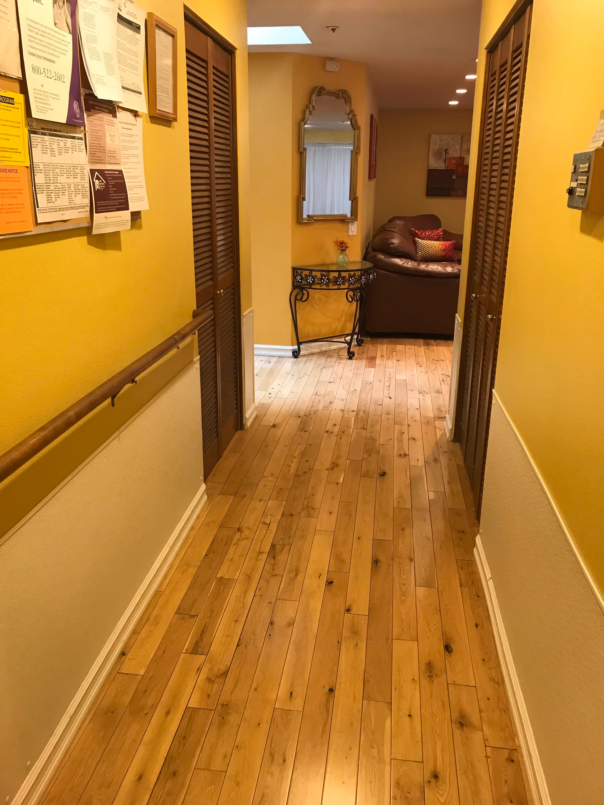 A hallway with wooden flooring and yellow walls. On the left wall, there is a bulletin board with various papers pinned to it and a wooden handrail below. On both sides of the hallway, there are wooden louvered doors. At the end of the hallway, there is a decorative table with a small vase of flowers and a mirror above it. Beyond the table, a brown leather couch with colorful pillows is partially visible in a living area.