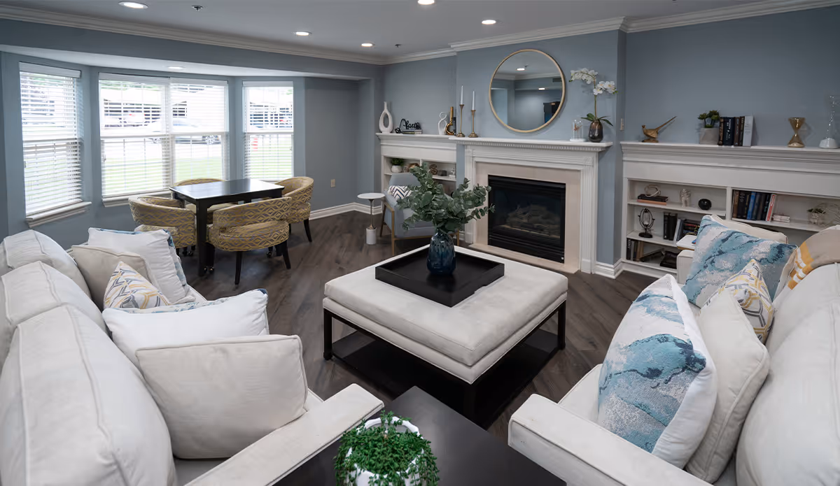 A cozy living room with light gray walls and dark wood flooring. The room features two beige sofas with decorative pillows, a large square ottoman with a black tray and a vase with greenery, a fireplace with a round mirror above it, built-in white shelves with books and decor, and a small dining table with four patterned chairs near a set of windows with white blinds.