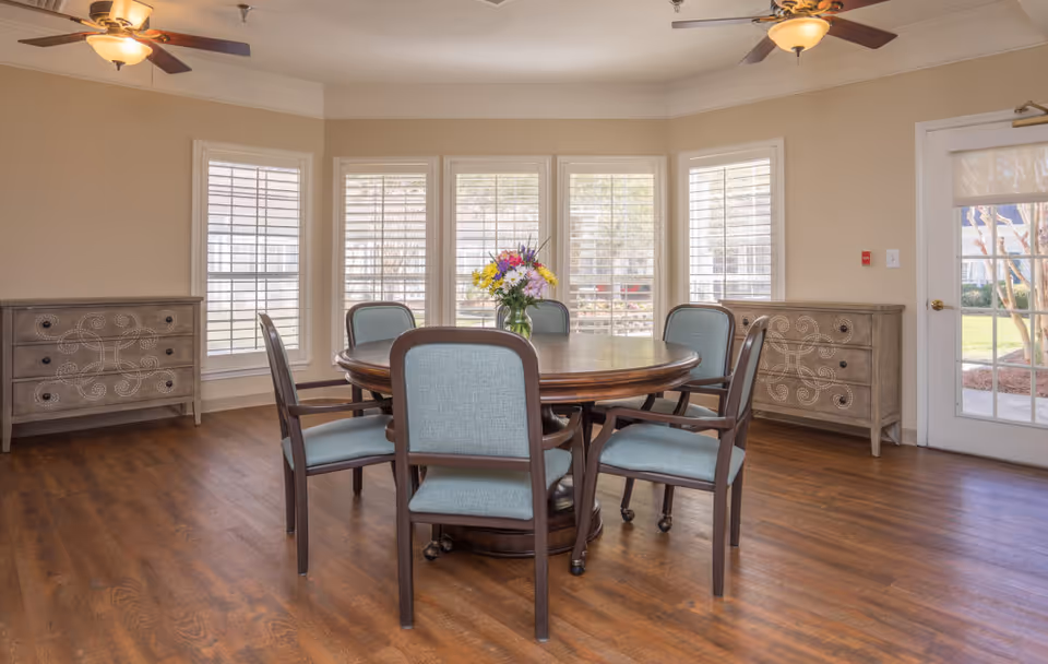 Bright communal dining room with a round wooden table and upholstered chairs, large shuttered windows, two decorative sideboards, and ceiling fans.